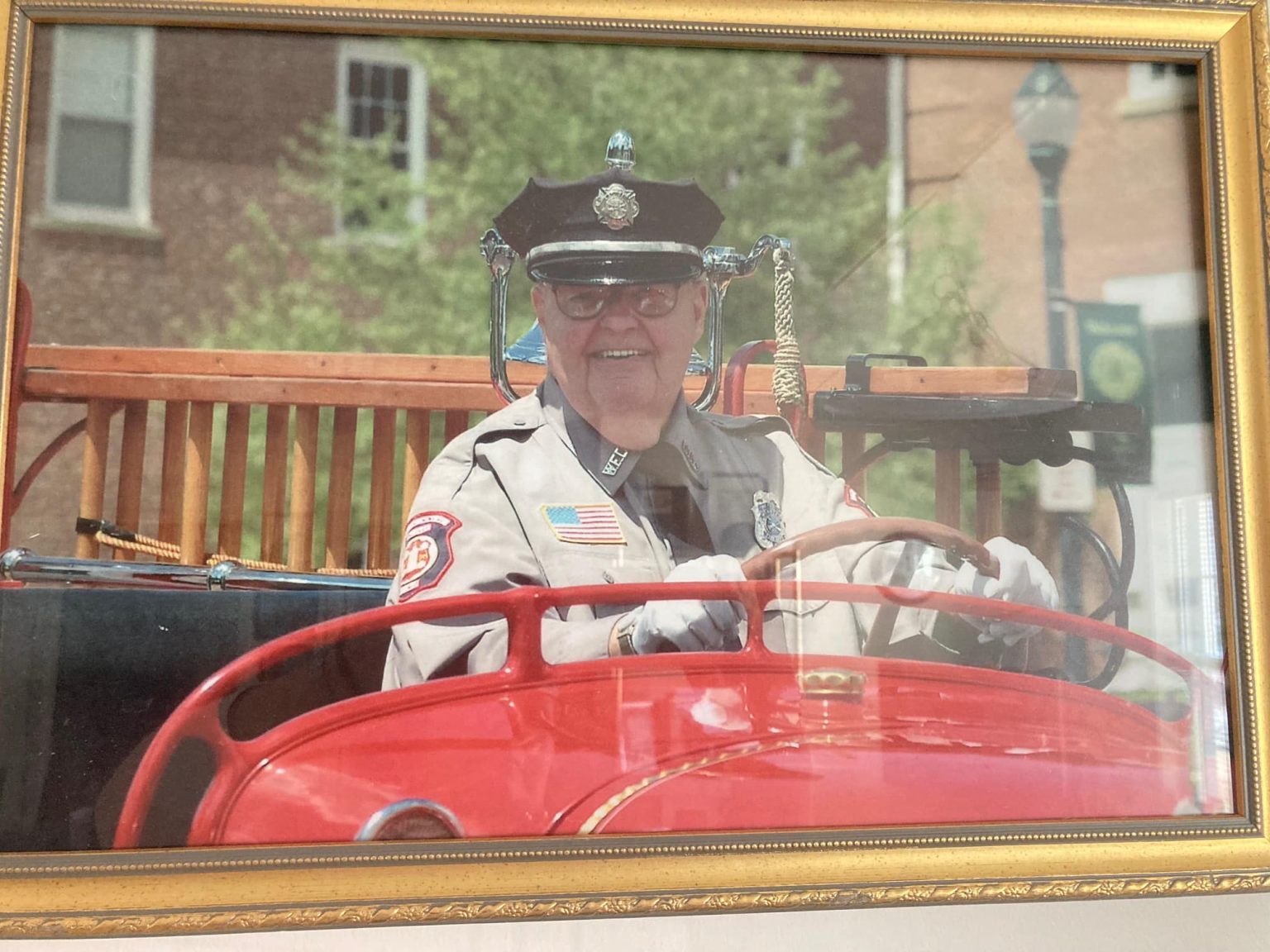 A smiling older person in a vintage police uniform drives a red antique fire truck. The truck is outdoors, and a brick building is visible in the background, framed by a window. The scene is captured within a gold-colored picture frame.