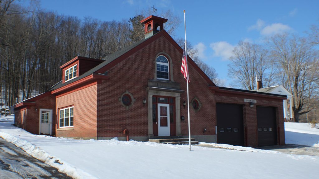 A brick fire station with two dark garage doors and a small cupola on the roof. An American flag is at half-mast in front. Snow covers the ground. Leafless trees are visible in the background under a clear blue sky.
