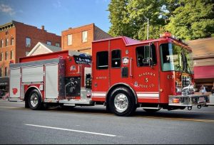 A red fire truck labeled "Niagara 3 Hose Co" is parked on a street. The vehicle features multiple compartments and equipment. In the background, there are brick buildings and trees under a blue sky.