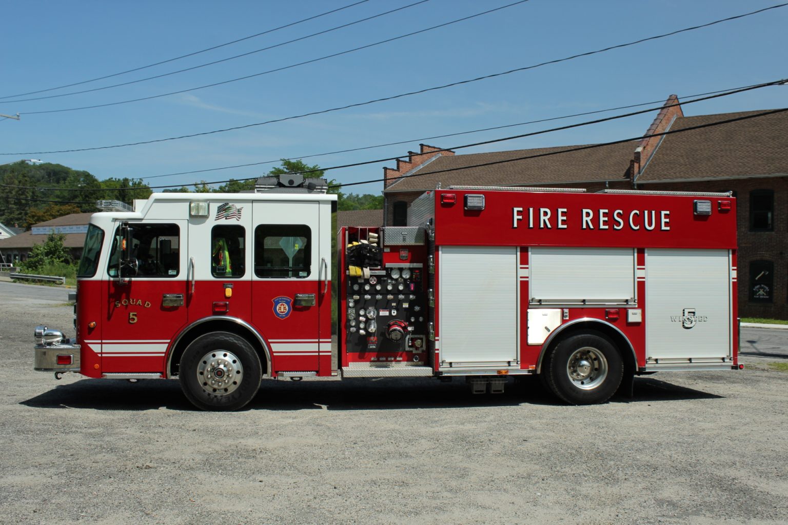 A red and white fire rescue truck is parked on a gravel lot in front of brick buildings, with overhead power lines visible in the background.