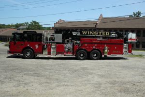A red fire truck labeled "Winsted" with an extended ladder is parked on a gravel area in front of a brick building and trees.