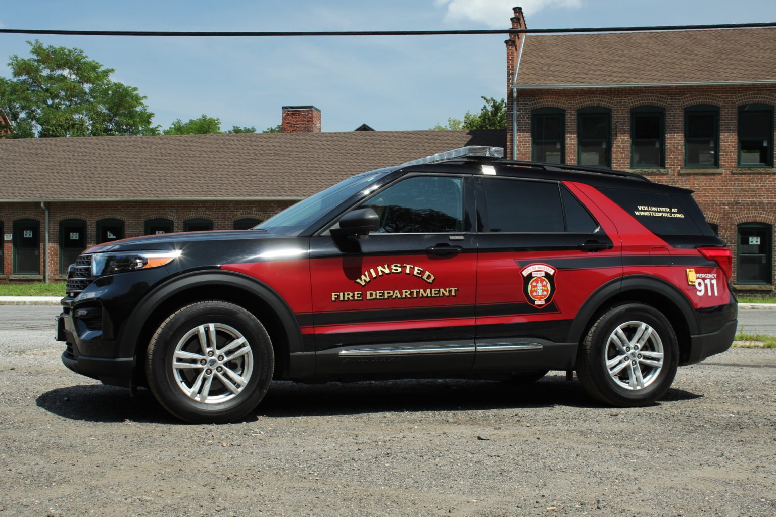 A black and red SUV labeled "Winsted Fire Department" with emergency and badge graphics is parked on gravel in front of a brick building with arched windows and a brown roof.