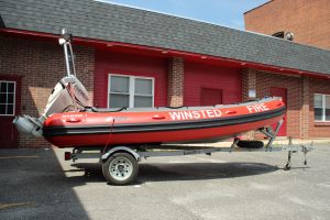 A red and black inflatable rescue boat labeled "WINSTED FIRE" is mounted on a trailer and parked outside a brick building with red doors and windows.