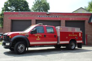 A red fire department tactical unit truck labeled "Winsted Tactical 8 Unit" and "Special Operations" is parked in front of a brick fire station with three garage doors.