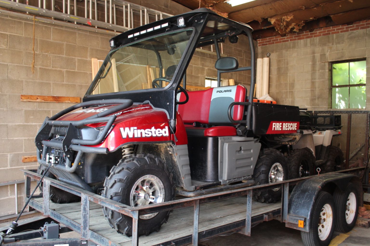 A red Polaris utility vehicle labeled "Winsted Fire Rescue" is parked on a trailer inside a garage with brick walls and ladders stored above.