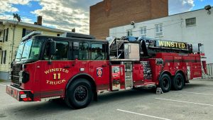 A red fire truck marked "Winsted Truck 11" is parked in a lot. The truck features a large ladder on top and various compartments along its side. Buildings and a cloudy sky are visible in the background.