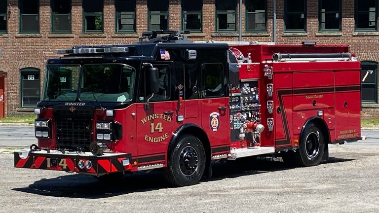 A red fire engine labeled "Winsted Engine 14" is parked on a gravel surface. The vehicle is equipped with various hoses and tools. Behind it is a brick building with green windows.