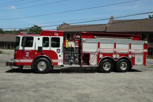 A red and white fire engine labeled "Pelham 2 Eng. Co." is parked on a gravel lot, with buildings and power lines visible in the background on a sunny day.