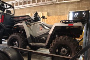 A gray all-terrain vehicle (ATV) with large tires and a police department emblem is loaded onto a trailer inside a garage with cinder block walls and tools visible in the background.