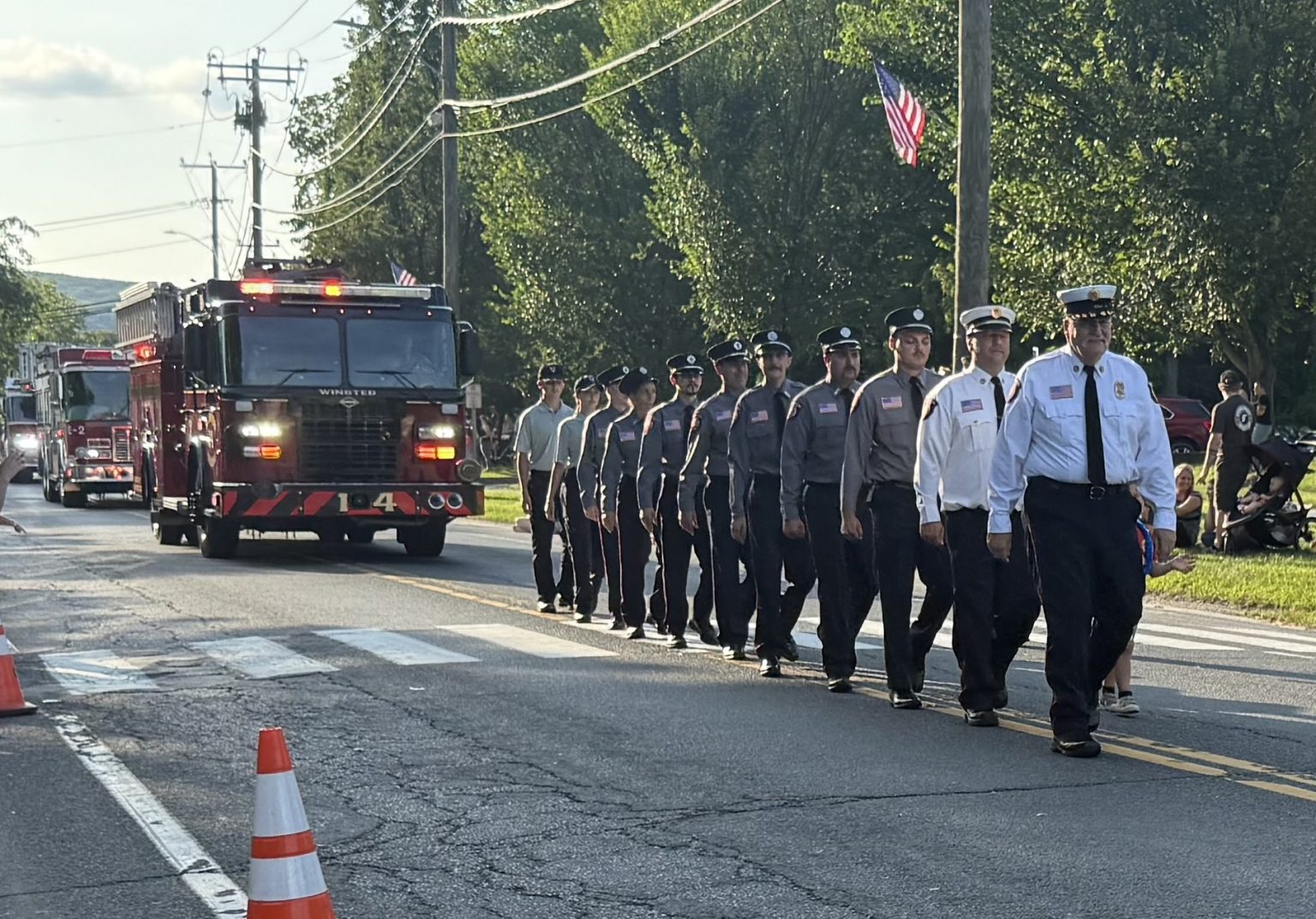 A group of firefighters in uniform march in formation down a sunlit street during a parade, with fire trucks behind them and people watching from the side. American flags and traffic cones are visible.