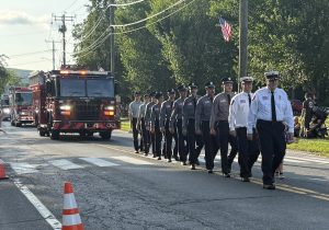 A group of firefighters in uniform march in formation down a sunlit street during a parade, with fire trucks behind them and people watching from the side. American flags and traffic cones are visible.