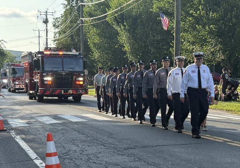 A group of firefighters in uniform march in formation down a sunlit street during a parade, with fire trucks behind them and people watching from the side. American flags and traffic cones are visible.